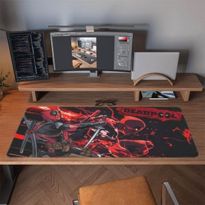 May include: A rectangular desk mat featuring a graphic illustration of the character Deadpool in red and black. The design shows the character in two poses with swords, set against a dark background with red accents and the text DEADPOOL in red.