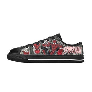 May include: A low-top sneaker featuring a black canvas upper with a comic book print of Spider-Man in red and blue. The shoe includes black laces, a black rubber toe cap, and a black sole with a thin white stripe running along the side.
