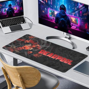 Könnte beinhalten: Eine rechteckige Schreibtischunterlage mit einer grafischen Illustration der Figur Deadpool in einem rot-schwarzen Anzug und dem Schriftzug DEADPOOL in fetten roten Buchstaben. Das Design umfasst ein dunkles, abstraktes Hintergrundmuster.