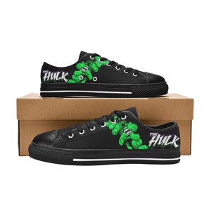 Puede incluir: Un par de zapatillas de lona negras de corte bajo con un estampado gráfico del personaje verde The Hulk en los laterales. Los zapatos tienen cordones negros, punteras de goma negra y suelas blancas con rayas negras. El texto The Hulk está impreso.