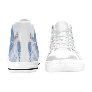 Pu&ograve; includere: Un paio di scarpe da ginnastica alte in tela bianca con una stampa grafica di un personaggio biondo in un lungo abito bianco e blu. Le scarpe hanno lacci bianchi, punte in gomma bianca e una sottile striscia nera lungo la base delle suole.