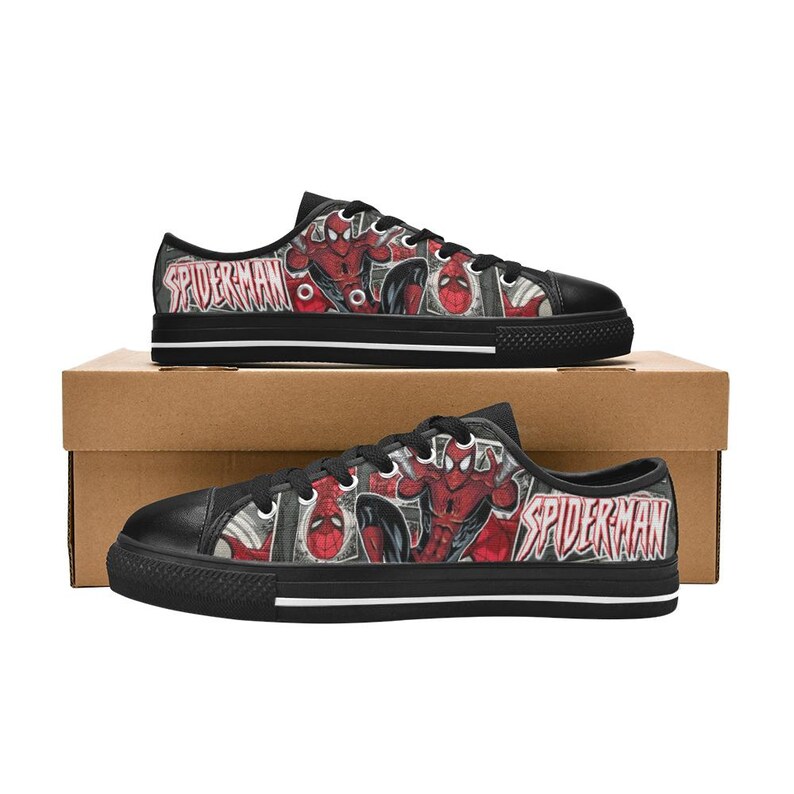 May include: Low-top sneakers featuring a black canvas upper with a red and black comic book print of Spider-Man and the text Spider-Man. The shoes have black laces, black rubber toe caps, and black soles with a thin white stripe along the side.