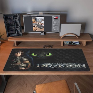 Può includere: Un tappetino da scrivania rettangolare nero con la scritta "HOW TO TRAIN YOUR DRAGON" e una grafica di un personaggio e degli occhi di drago. Il tappetino è su una scrivania di legno con monitor di computer.