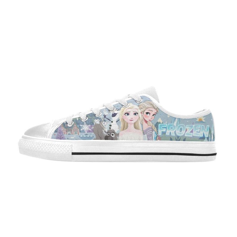 May include: A low-top canvas sneaker featuring a light blue and white winter-themed print with characters from Frozen, including Anna, Elsa, Olaf, and Sven. The shoe has white laces, a white rubber toe cap, and a white sole with a thin black stripe.