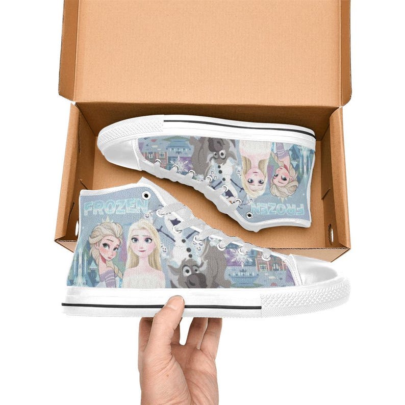 Pode incluir: Um par de t&eacute;nis de cano alto em lona branca com um gr&aacute;fico impresso de personagens do filme Frozen, incluindo Elsa, Anna e Sven. Os sapatos t&ecirc;m atacadores brancos, biqueiras de borracha branca e solas brancas dentro de uma caixa de cart&atilde;o.