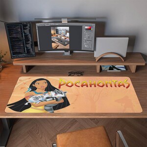 Puede incluir: Alfombrilla de escritorio rectangular con una imagen de dibujos animados de Pocahontas sosteniendo un mapache. La alfombrilla tiene un fondo beige con la palabra "POCAHONTAS" en naranja. La alfombrilla está sobre un escritorio de madera con un monitor de computadora, una computadora portátil y otros suministros de oficina.