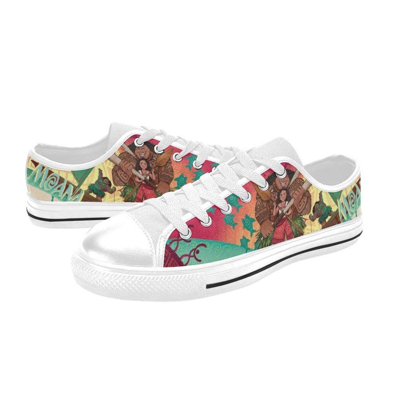 May include: White canvas sneakers with a colorful design featuring characters and the word "Moana". The shoes have white laces, a white rubber toe cap, and a black stripe along the sole. The design includes shades of red, orange, green, and brown.