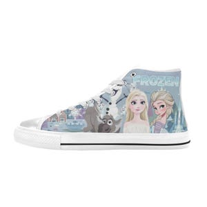 Pode incluir: Um t&ecirc;nis de cano alto com fundo azul claro e a palavra FROZEN impressa em branco. O design inclui ilustra&ccedil;&otilde;es das personagens Elsa, Anna, Olaf e Sven, juntamente com padr&otilde;es de flocos de neve e a silhueta de um castelo.