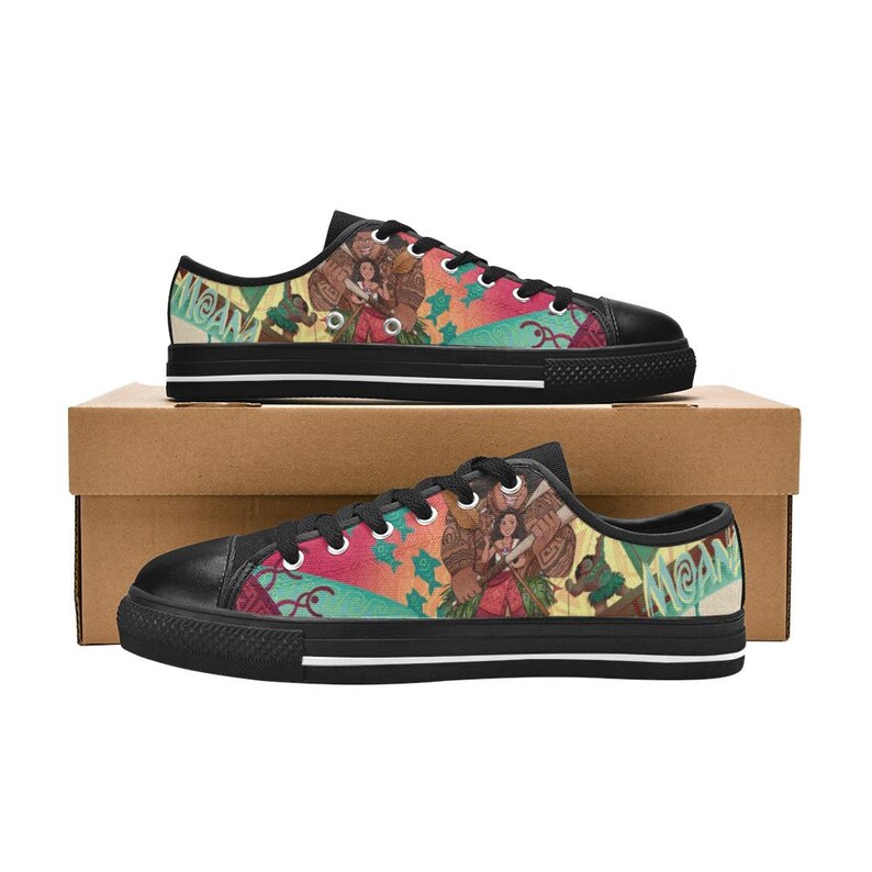 May include: Black canvas sneakers with a colorful design featuring characters and the word "MOANA". The shoes have black laces, soles, and toe caps. The design includes shades of red, green, and blue.