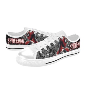 May include: White canvas sneakers featuring a Spider-Man design. The shoes have a white toe cap, white laces, and a black stripe along the sole. The Spider-Man graphic is red and black, with the words "SPIDER-MAN" in red.