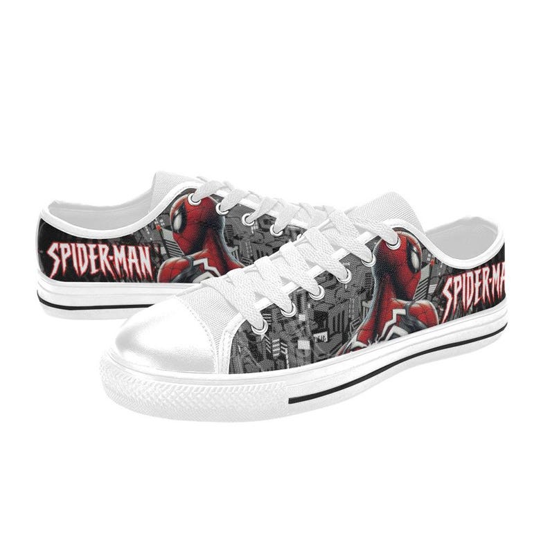 May include: White canvas sneakers featuring a Spider-Man design. The shoes have a white toe cap, white laces, and a black stripe along the sole. The Spider-Man graphic is red and black, with the words "SPIDER-MAN" in red.