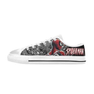 May include: White canvas low-top sneakers featuring a Spider-Man design. The sneakers have a black and white cityscape pattern on the side and the Spider-Man logo on the heel. The Spider-Man character is depicted in red and black.