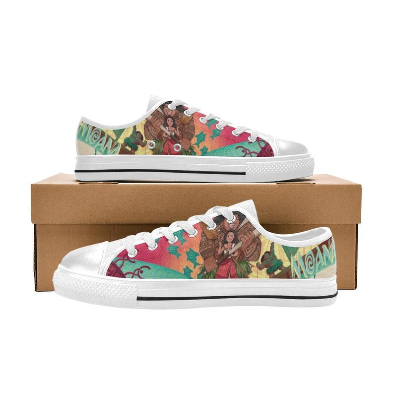 May include: White canvas sneakers featuring a colorful design with characters and the word "MOANA". The shoes have a white rubber sole and laces. The design includes shades of red, green, and blue.