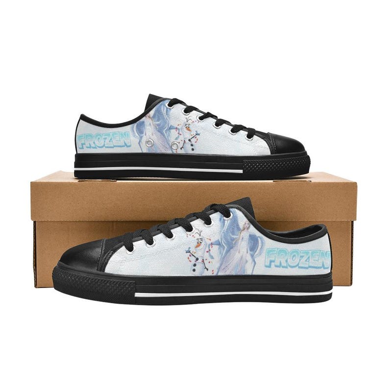 Peut inclure: Baskets basses en toile avec une base blanche et une impression graphique d'Elsa et Olaf de Frozen. Les chaussures ont des bouts en caoutchouc noir, des lacets noirs, une bordure noire et une semelle interm&eacute;diaire blanche avec une bande noire.