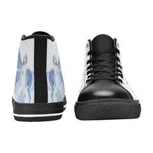 Pu&ograve; includere: Un paio di scarpe alte con tomaia in tela bianca caratterizzata da un'illustrazione ripetuta di un personaggio biondo con un lungo abito bianco e dettagli blu. Le scarpe presentano lacci neri, punte in gomma nera e suole nere con una riga bianca.