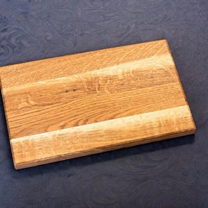 Puede incluir: Una tabla de cortar rectangular de madera clara, con vetas visibles. Está hecha de varias tablas unidas. El fondo es una tela azul oscuro con un patrón sutil.