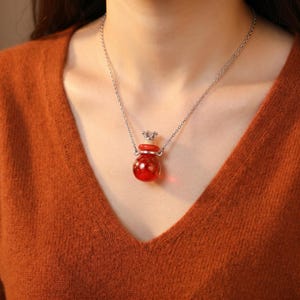 Op de afbeelding: Een zilveren ketting met een kleine, ronde, rode glazen fleshanger. De fles heeft een zilveren kroonvormige stopper. De ketting wordt gedragen op een roestkleurige trui.