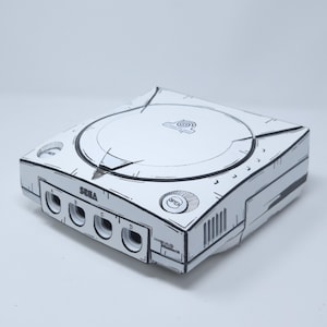 Può includere: Una console per videogiochi Sega Dreamcast bianca con un'estetica cel-shaded disegnata a mano e contorni neri marcati. La console include quattro porte per controller etichettate A, B, C e D, un coperchio circolare per il disco e pulsanti di accensione.