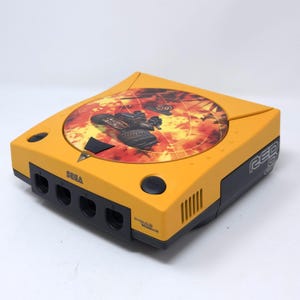 Może przedstawiać: Żółta konsola do gier wideo Sega Dreamcast z okrągłą grafiką na górnej pokrywie przedstawiającą pojazd w ognistym krajobrazie. Konsola posiada cztery porty kontrolerów z przodu, czarny przycisk otwierania oraz napis Red 0 na bocznej stronie.