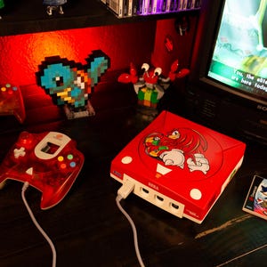 Puede incluir: Una consola de videojuegos Sega Dreamcast roja con un gráfico de Knuckles the Echidna en la tapa superior, junto a un mando con cable rojo translúcido y una caja del juego Sonic Adventure 2, situada cerca de una pantalla de televisión con texto.