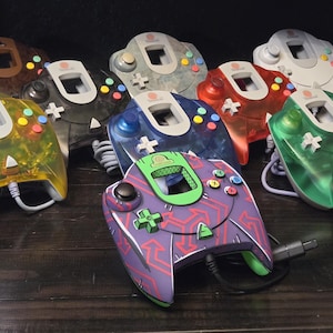 Könnte beinhalten: Eine Sammlung von Sega Dreamcast-Gamecontrollern in verschiedenen Farben und Ausführungen, darunter durchscheinendes Gelb, Schwarz, Blau, Orange und Grün, ein einfarbiges Weiß, ein Holzmaserungsmuster und ein lila Controller mit grünem Pfeilmotiv.
