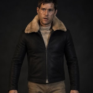 May include: A black leather jacket with a tan shearling collar. The jacket has a zip closure and is worn over a light-coloured sweater. The person is wearing tan trousers. The background is dark.