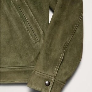 Puede incluir: Primer plano de una chaqueta de ante verde oliva. La chaqueta presenta un bolsillo delantero, pu&ntilde;os abotonados y detalles de costura blancos. La textura suave y el color sugieren una prenda de abrigo informal pero elegante.