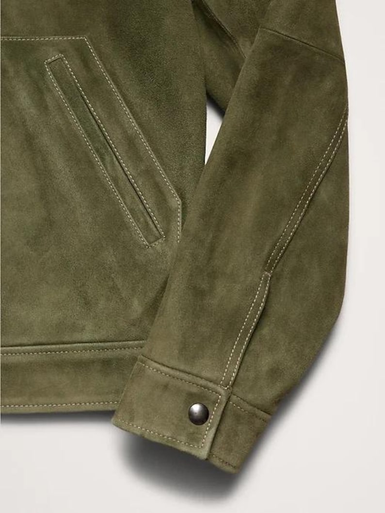 Puede incluir: Primer plano de una chaqueta de ante verde oliva. La chaqueta presenta un bolsillo delantero, pu&ntilde;os abotonados y detalles de costura blancos. La textura suave y el color sugieren una prenda de abrigo informal pero elegante.