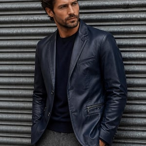 May include: A navy blue leather blazer with a two-button closure and a zippered chest pocket. The blazer is paired with a dark blue sweater and gray trousers. The model is standing in front of a gray metal background.
