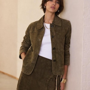 Puede incluir: Chaqueta de ante verde oliva con falda cruzada a juego. La chaqueta tiene cuello clásico, cierre de botones y dos bolsillos delanteros. La falda tiene un diseño cruzado con cierre de lazo. El conjunto se combina con una camiseta blanca y joyas doradas.