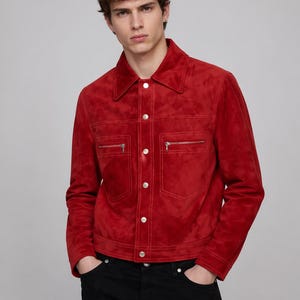 May include: A red suede jacket with a classic collar, two zipped chest pockets, and silver buttons. The jacket has a tailored fit and long sleeves. The model is wearing black jeans.