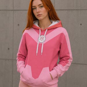 Puede incluir: Una sudadera con capucha rosa con forro de capucha blanco y cordones blancos. La sudadera presenta un diseño geométrico en tonos rosas y un emblema circular en el pecho. La modelo lleva pantalones cortos rosas y tiene el pelo rojo.