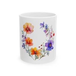 Może przedstawiać: Biały ceramiczny kubek z akwarelowym wzorem kwiatowym. Wzór zawiera wieniec kolorowych kwiatów w odcieniach pomarańczu, różu, fioletu i niebieskiego. Kwiaty rozmieszczone są wokół zewnętrznej strony kubka.