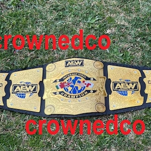 May include: A gold and black wrestling championship belt with the text "AEW International Champion". The belt has gold plates with intricate designs and the AEW logo. The belt is lying on a bed of green grass.