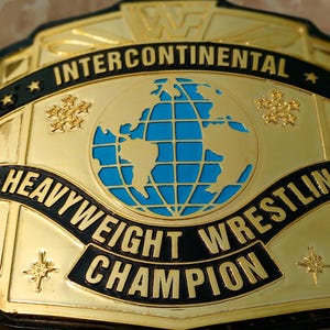 May include: A gold-colored wrestling championship belt with black and blue accents. The belt features the words "Intercontinental Heavyweight Wrestling Champion" and a blue globe design. The side plates have a gold emblem.