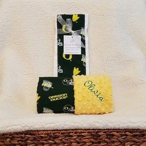 May include: A set of baby items, including a green and yellow patterned blanket, a small square item with the name "Olivia" embroidered on it, and a matching green and yellow patterned item. The items are tied with a white ribbon.