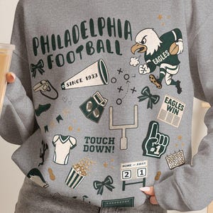 May include: A heather grey crewneck sweatshirt featuring a graphic print with the text Philadelphia Football, an eagle mascot, a megaphone, a goalpost, a foam finger, a scoreboard, and various football-themed icons in dark green, white, and tan.