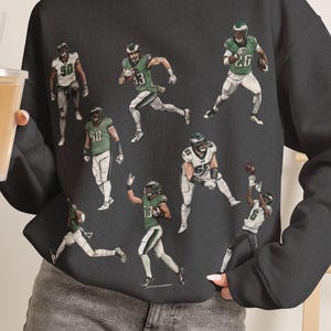 Puede incluir: Una sudadera de cuello redondo en color gris carbón con un estampado gráfico repetitivo de jugadores de fútbol americano con uniformes verdes y blancos en varias poses de acción, incluyendo correr, atrapar y lanzar un balón, con mangas largas.