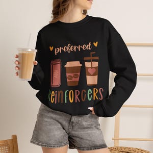 May include: A black long-sleeve sweatshirt featuring a graphic print with the text "preferred REINFORCERS" in colorful letters. The design includes illustrations of a soda can, a coffee cup, and a tall iced coffee drink with a heart icon.
