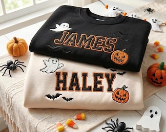 Puede incluir: Una sudadera negra con el nombre JAMES bordado en naranja y una sudadera beige con el nombre HALEY bordado en negro. Ambas presentan fantasmas, calabazas y murciélagos bordados. Las sudaderas están apiladas sobre una mesa.