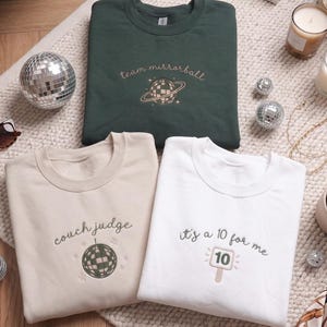 May include: Three crewneck sweatshirts in dark green, beige, and white. The dark green sweatshirt features an embroidered planet and the text team mirrorball. The beige sweatshirt shows a disco ball and the text couch judge. The white sweatshirt displays a score paddle with the number 10 and the text it's a 10 for me.