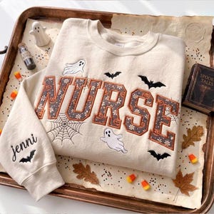 Puede incluir: Una sudadera de cuello redondo color crema con la palabra NURSE en letras grandes con un patrón oscuro de flores y calabazas. El diseño incluye fantasmas blancos bordados, murciélagos negros y una telaraña. La manga tiene bordado el nombre jenni.