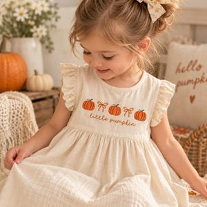 Pode incluir: Um vestido sem mangas na cor creme com ombros franzidos, apresentando um design bordado de três abóboras laranja e dois laços bege, com o texto "little pumpkin" costurado em fio castanho na zona do peito.