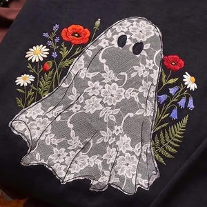 Puede incluir: Una sudadera negra con un diseño bordado de un fantasma de encaje blanco. El fantasma está rodeado de coloridas flores silvestres bordadas, incluyendo amapolas rojas, margaritas blancas y flores azules en forma de campana con tallos verdes.