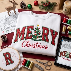 May include: A white sweatshirt featuring an embroidered design with the text MERRY CHRISTMAS in red glittery letters, where the letter E is replaced by a green Christmas tree graphic. The design includes red and green thread spools.