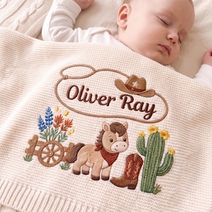 May include: A cream-colored knit baby blanket featuring an embroidered design of a brown pony, a cowboy boot, a cactus, a wooden wagon wheel, and flowers. The name Oliver Ray is stitched in brown letters inside a lasso-shaped border.
