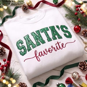 May include: A white crewneck sweatshirt featuring the text SANTA'S in green glittery block letters and the word favorite in red cursive script. The garment has a ribbed collar and long sleeves, showcasing a clean and simple design.