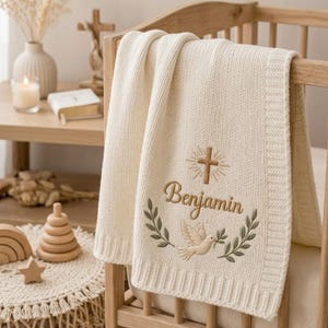 May include: A cream-colored knitted baby blanket featuring an embroidered design of a brown cross, the name Benjamin in brown script, and a dove surrounded by green olive branches, draped over the side of a wooden crib.