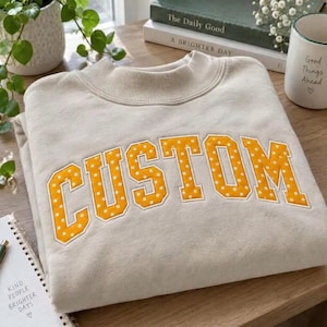 Può includere: Una felpa girocollo beige con la scritta CUSTOM in grandi lettere di tessuto giallo a pois applicate sul petto. La felpa presenta un colletto a coste e una consistenza morbida, proposta in una tonalità neutra.