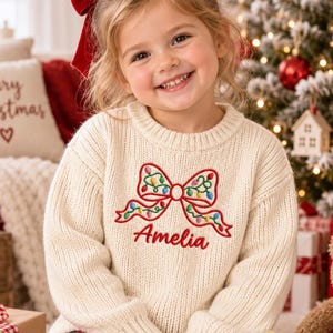 May include: A cream-colored, long-sleeved, ribbed knit sweater featuring an embroidered red bow design decorated with small, colorful Christmas light bulbs. The name Amelia is embroidered in red script lettering directly below the bow.